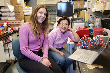 UCLA Graduate Student Noelle Mitchell wearing a pink sweater (left), and Dr. Kyungae Yang of Columbia in a pink button-up shirt (right), smiling in a research lab with equipment all around them.