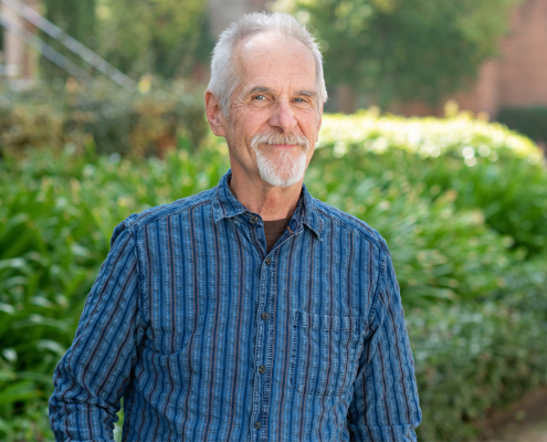 Paul Kroskrity smiling in a black-and-blue long-sleeve collar shirt, right hand in his pocket, left hand hanging at this side, with brick buildings and green plants and trees in the background.