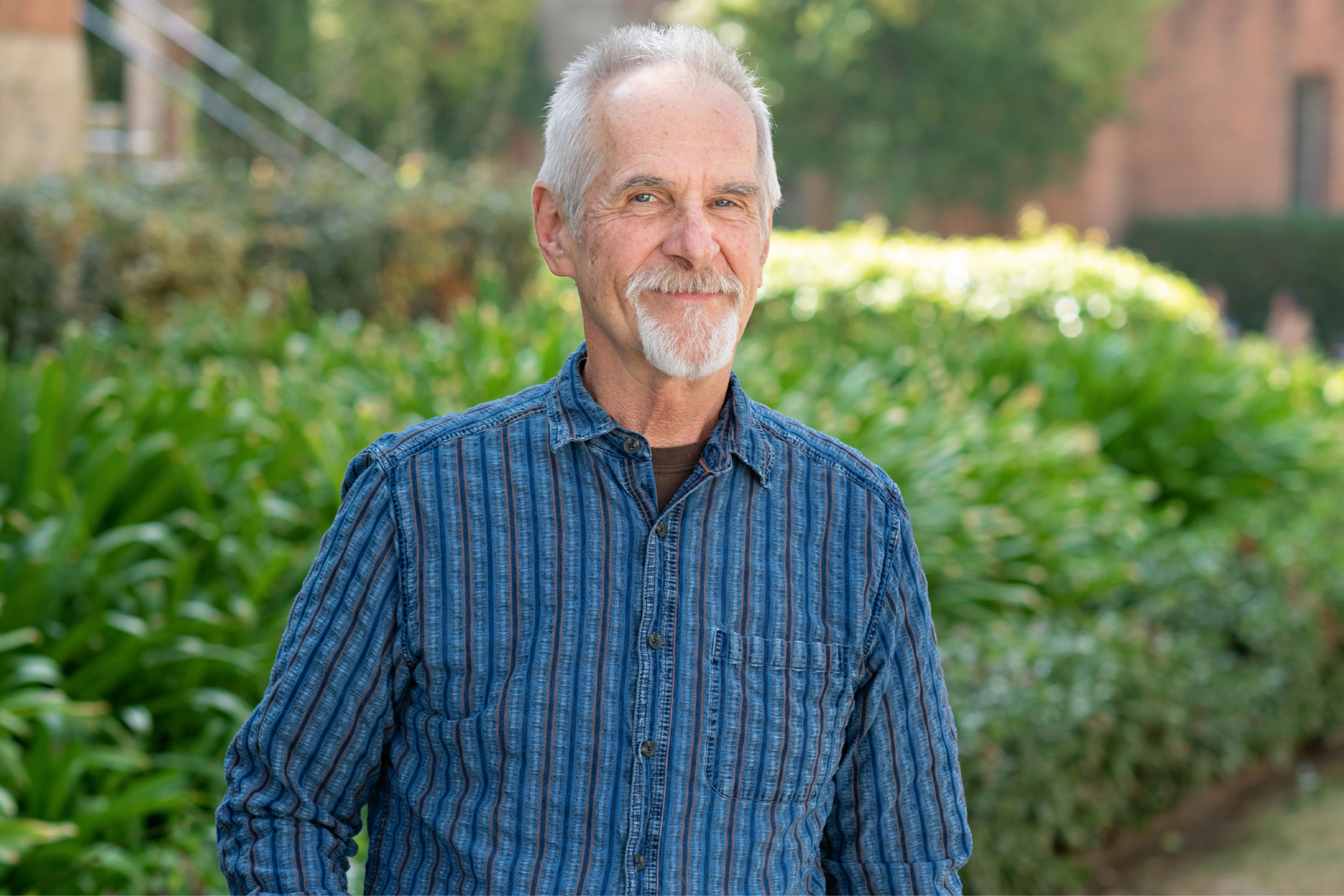 Paul Kroskrity smiling in a black-and-blue long-sleeve collar shirt, right hand in his pocket, left hand hanging at this side, with brick buildings and green plants and trees in the background.