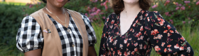 Tiffany Brannon (left) smiles in a black-and-white checkered dress and tan, sleeveless overcoat, hands clasped before her; Riley Marshall (right) smiles in a black floral dress, left hand on her waist; green foliage, pink flowers and the arches of Royce Hall stand behind them.