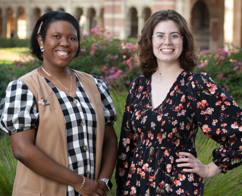 Tiffany Brannon (left) smiles in a black-and-white checkered dress and tan, sleeveless overcoat, hands clasped before her; Riley Marshall (right) smiles in a black floral dress, left hand on her waist; green foliage, pink flowers and the arches of Royce Hall stand behind them.