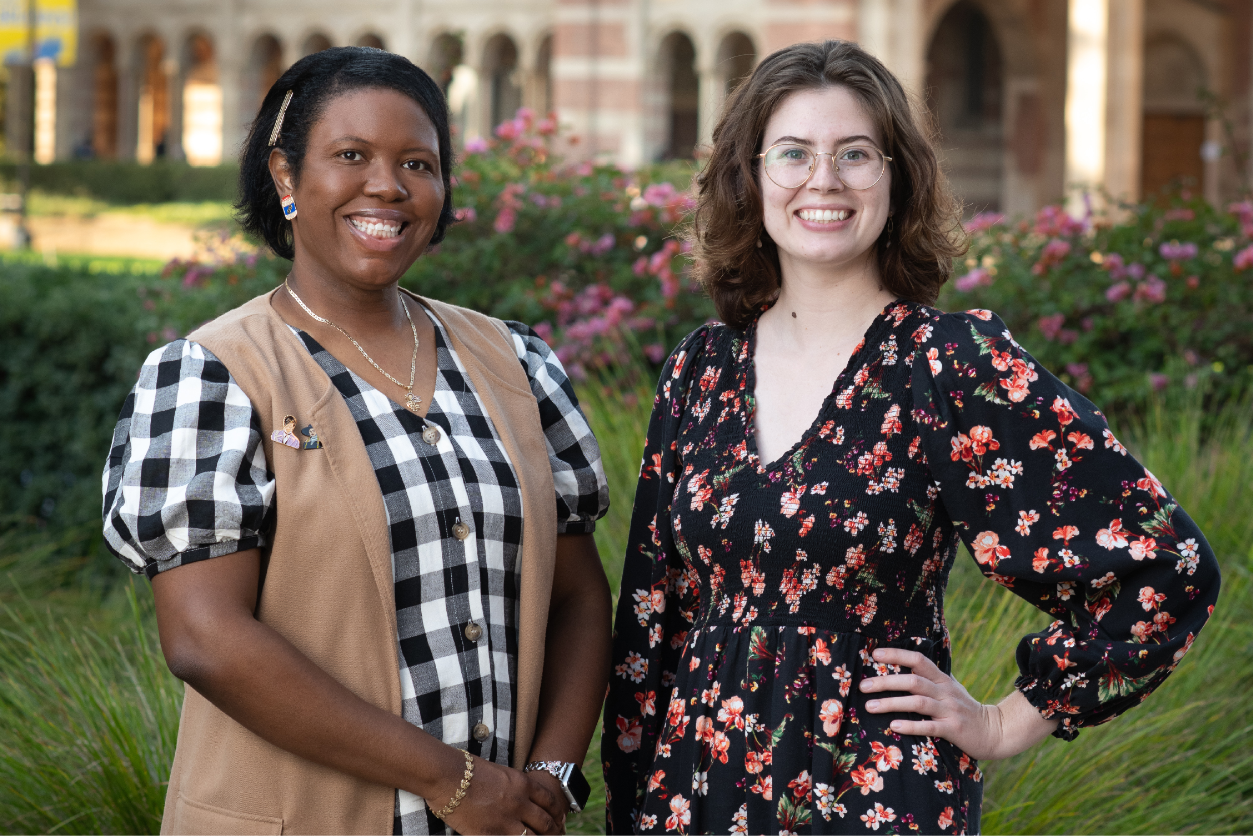 Tiffany Brannon (left) smiles in a black-and-white checkered dress and tan, sleeveless overcoat, hands clasped before her; Riley Marshall (right) smiles in a black floral dress, left hand on her waist; green foliage, pink flowers and the arches of Royce Hall stand behind them.