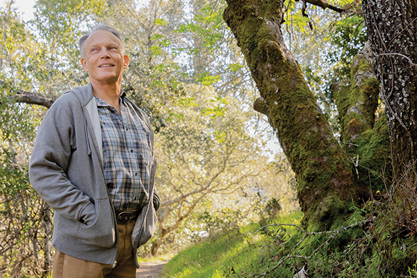 Hollis Lenderking on a hiking trail wearing hiking boots, khaki pants, and a collared shirt and grey sweater, both hands inside his sweater pockets, surrounded by trees and a blue sky in the background.