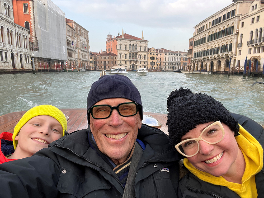 From left to right, Teddy, Peter and Sheri Weller on a boat with water and buildings in the background of a grey-blue sky. 