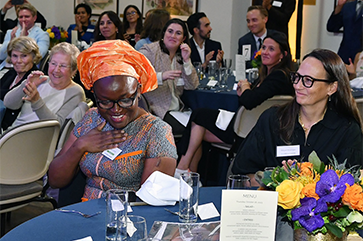 Iroro Tanshi, in orange head covering, sits at dining table in room full of people and holds hand to her chest