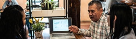 Three Latinos of varying ages sitting at a table with a laptop, pad of paper, phone and tablet