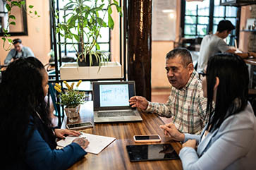 Three Latinos of varying ages sitting at a table with a laptop, pad of paper, phone and tablet