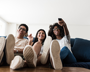 Teenagers sitting close to each other on sofa with their feet on a coffee table; one is holding a TV remote