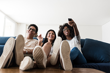 Teenagers sitting close to each other on sofa with their feet on a coffee table; one is holding a TV remote