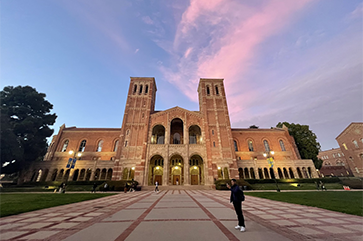 Wide shot of Royce Hall with blue sky and pink clouds, man looking at phone in foreground