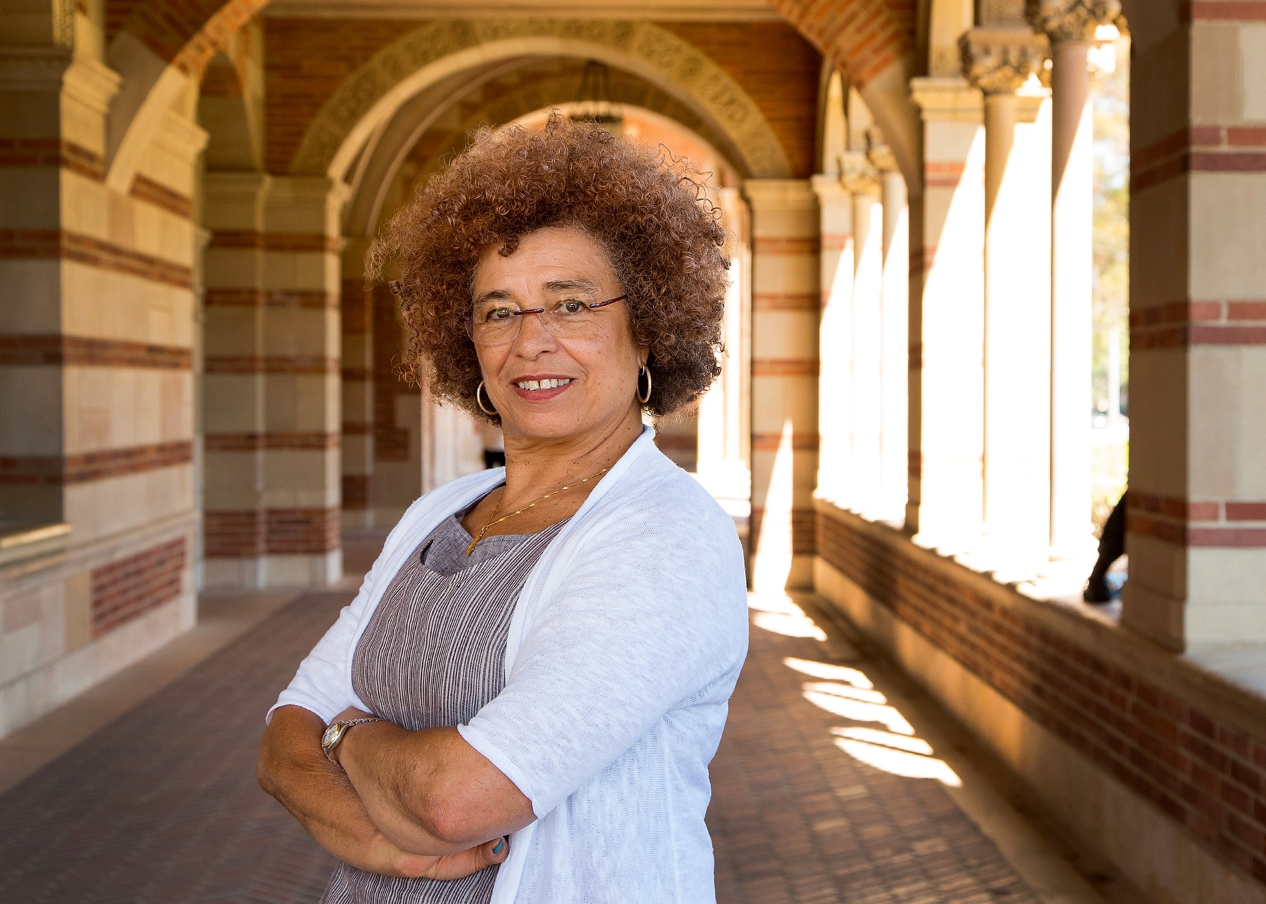Angela David posing with her arms crossed as she stands in a brick, stone and terracotta corridor, with the sun illuminating stone columns in the background.