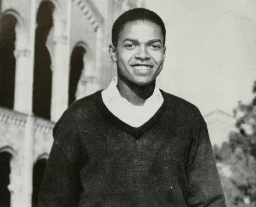 A black and white photograph of Sherrill Luke smiling, wearing a black sweater with a white college underneath, and a building with columns in the background.