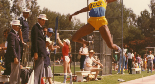 Jackie Joyner-Kersee in blue and gold UCLA exercise clothing performing a long distance jump with spectators in the background.