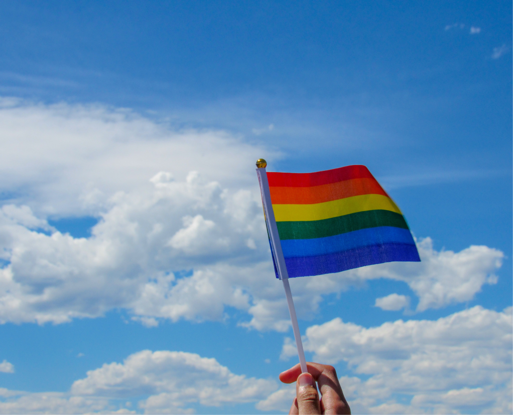 An image of someone holding a miniature rainbow flag or pride flag with clouds and a blue sky in the background.
