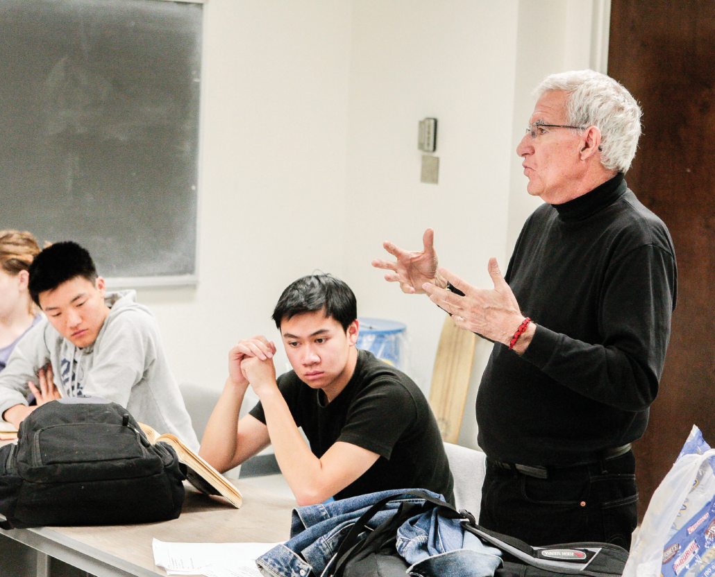 A male professor lecturing in a class room with a chalkboard with two male students listening attentively.