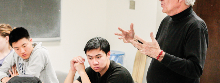 A male professor lecturing in a class room with a chalkboard with two male students listening attentively.