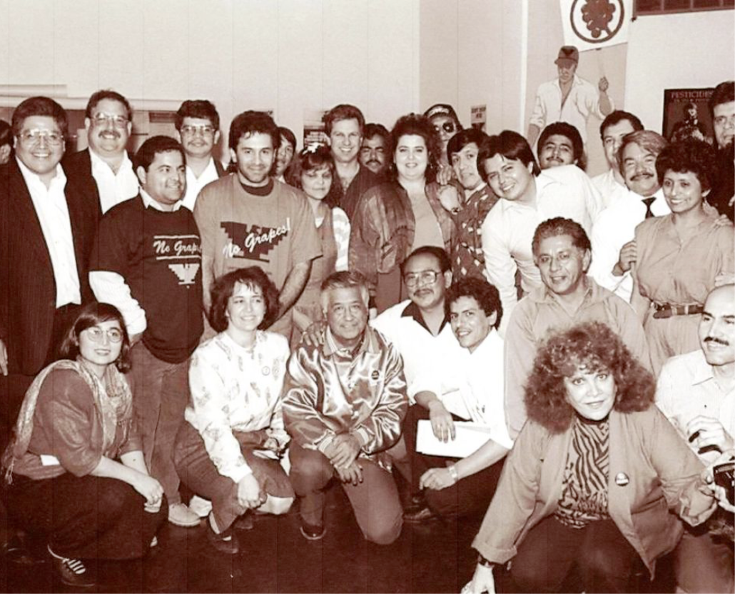 A black and white photograph of César E. Chávez surrounded twenty plus people as they pose for the photograph.