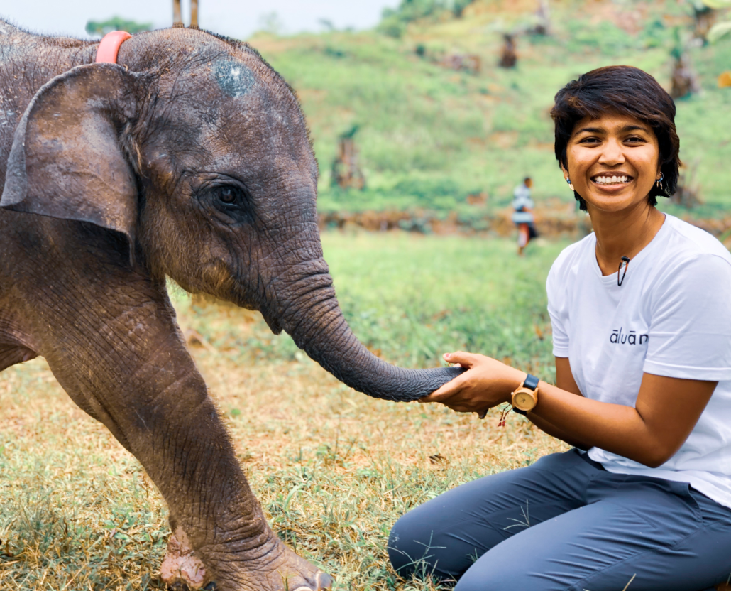 A woman wearing grey pants and a white shirt in an outdoor setting resting on her knees as she plays with a baby elephant.
