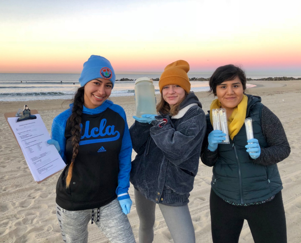 Three females students with lab equipment standing by a sandy beach, with waves and a multicolored sky in the background.