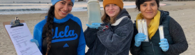 Three females students with lab equipment standing by a sandy beach, with waves and a multicolored sky in the background.
