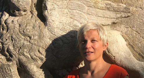Zrinka Stahuljak in a red shirt standing in front of stone griffin whose face has eroded.