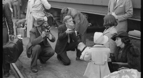 A black and white photograph of cameramen film children during a forced removal proceeding of Japanese Americans in the U.S. during WWII in the 1940s.