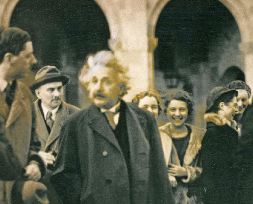 A black and white photograph depicting Albert Einstein surrounded by men in business attire with young female students smiling in the background.