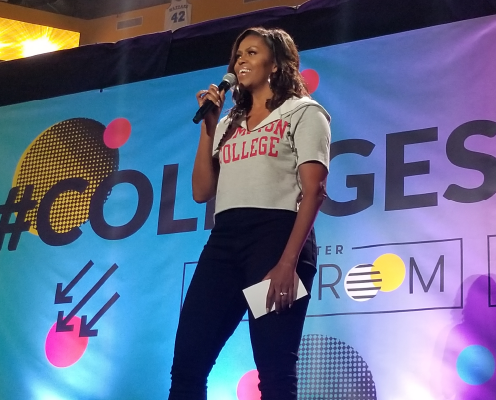 Michelle Obama speaking with a microphone in her hand, with a blue and purple banner in the background.