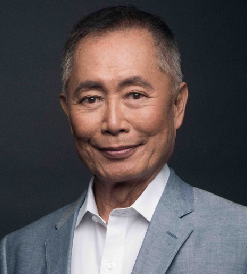 A head and shoulders photograph of George Takei in a grey suit against a dark grey background.