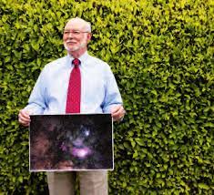 Fred Eiserling wearing a dress shirt with a red tie holding a photograph depicting a galaxy and stars, with a tall verdant shrub in the background.