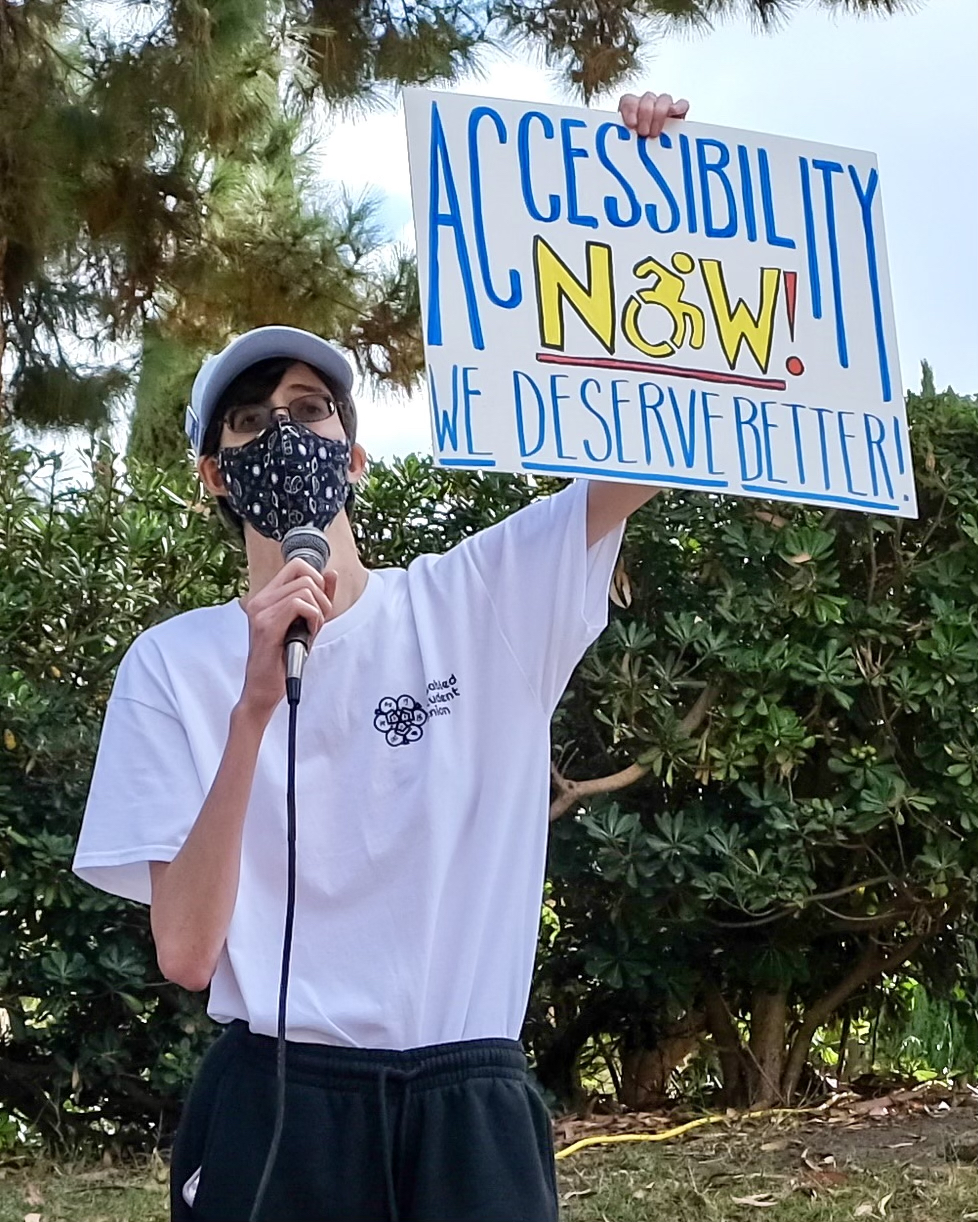 Christopher Ikonomou wearing a black cloth mask holding a microphone and a white poster that reads "Accessibility NOW! We deserve better!"
