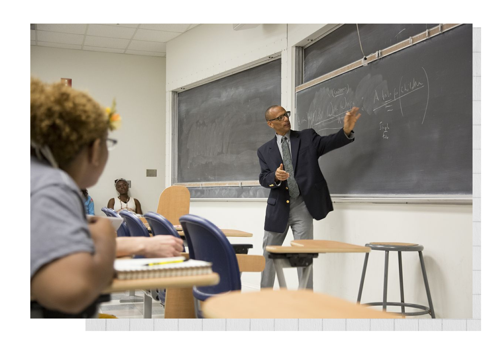 Professor Fred D’Aguiar in a suit and tie teaching to students in a classroom with a chalkboard.