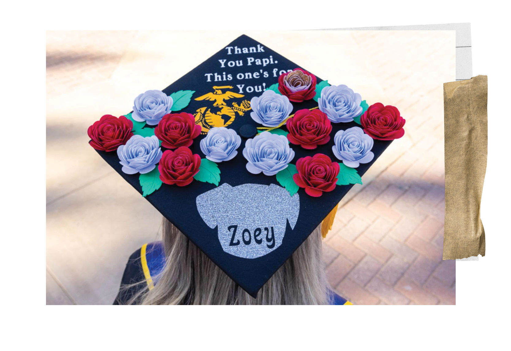 A student wearing a graduation cap decorated with light blue and red roses, green leaves, a paper-cut out of a dog with the name "Zoey," stenciled atop, the Marine Corps.' Eagle, Globe, an Anchor and the words, "Thank you, Papi. This one's for you!"
