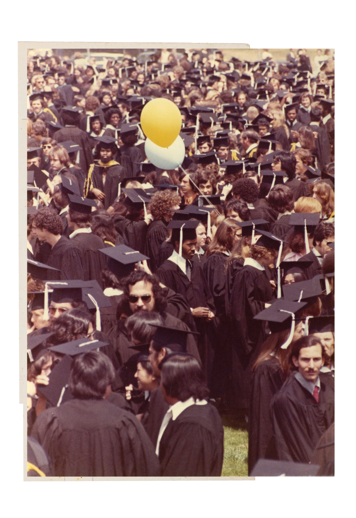 Students from the 1975 UCLA College graduation ceremony dressed in caps and gowns, with a white and yellow balloon floating overheard.
