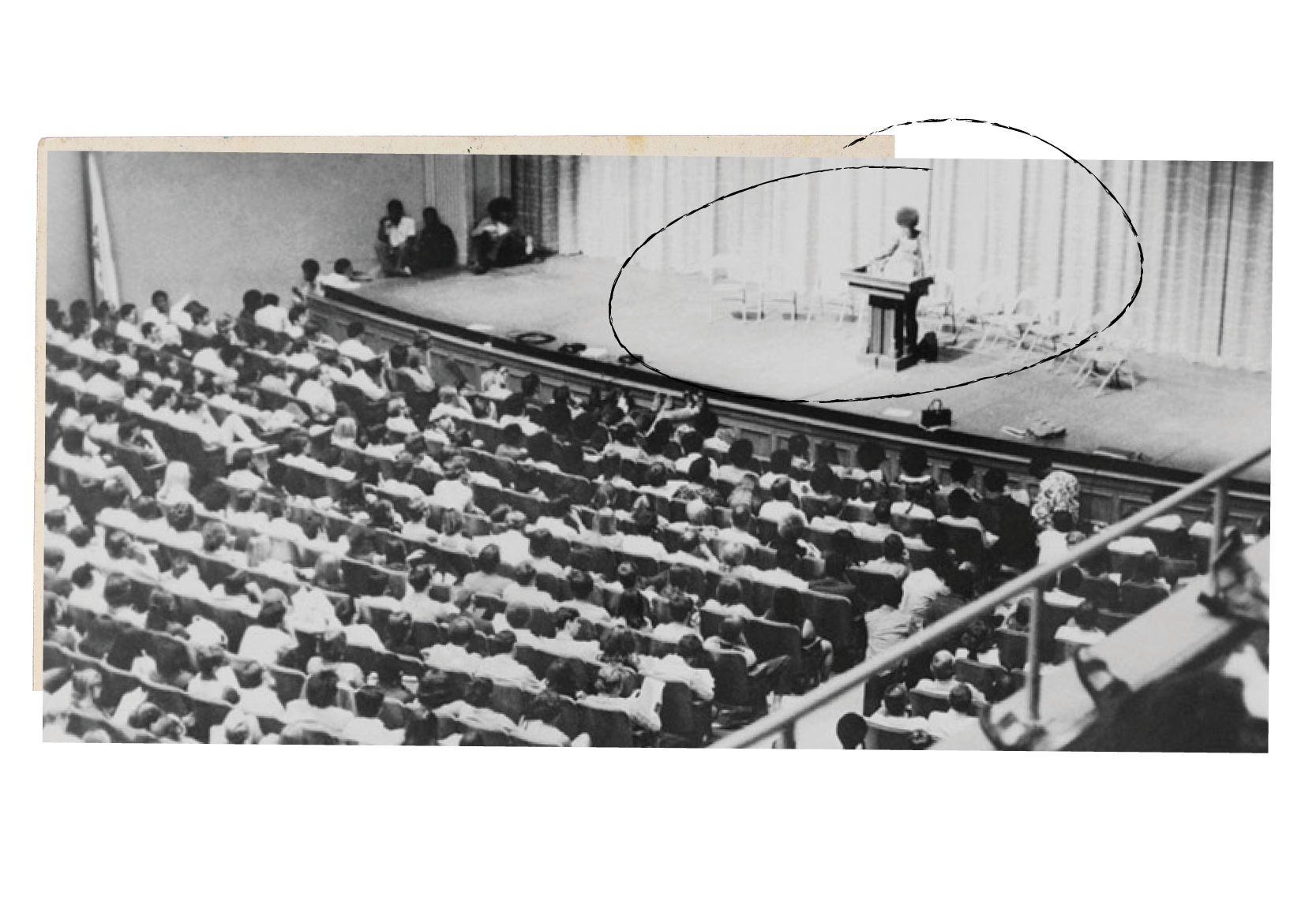 A black and white photograph of Angela Davis standing at a podium as she delivers a lecture to students in a lecture hall. ﻿