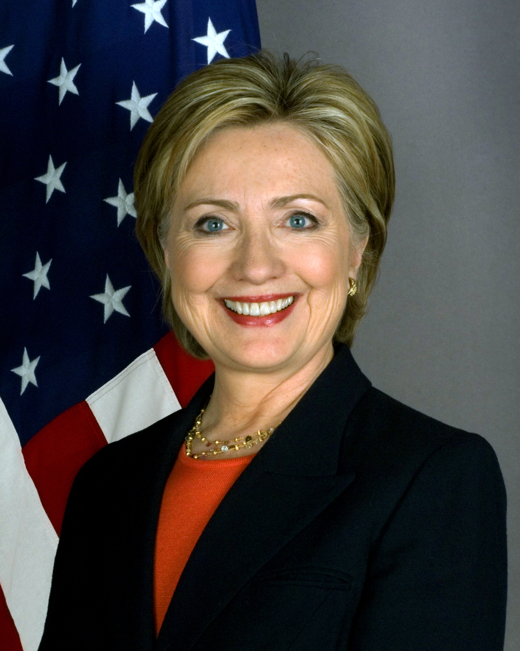 Hillary Clinton in a black sport coat with a red dress shirt underneath, with the United States flag in the background.