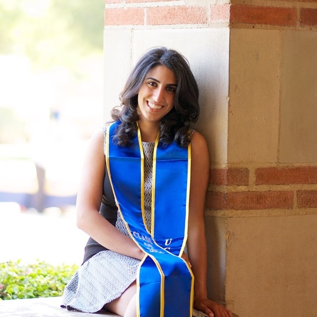 Jessica Kianmahd sitting on a concrete bench whilst leaning on a concrete and red brick column, wearing a blue graduation sash adorned with yellow accents.