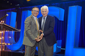 Tony Pritzker and Chancellor Block stand together holding Fiat Lux statue in front of blue block letters reading “UCLA”