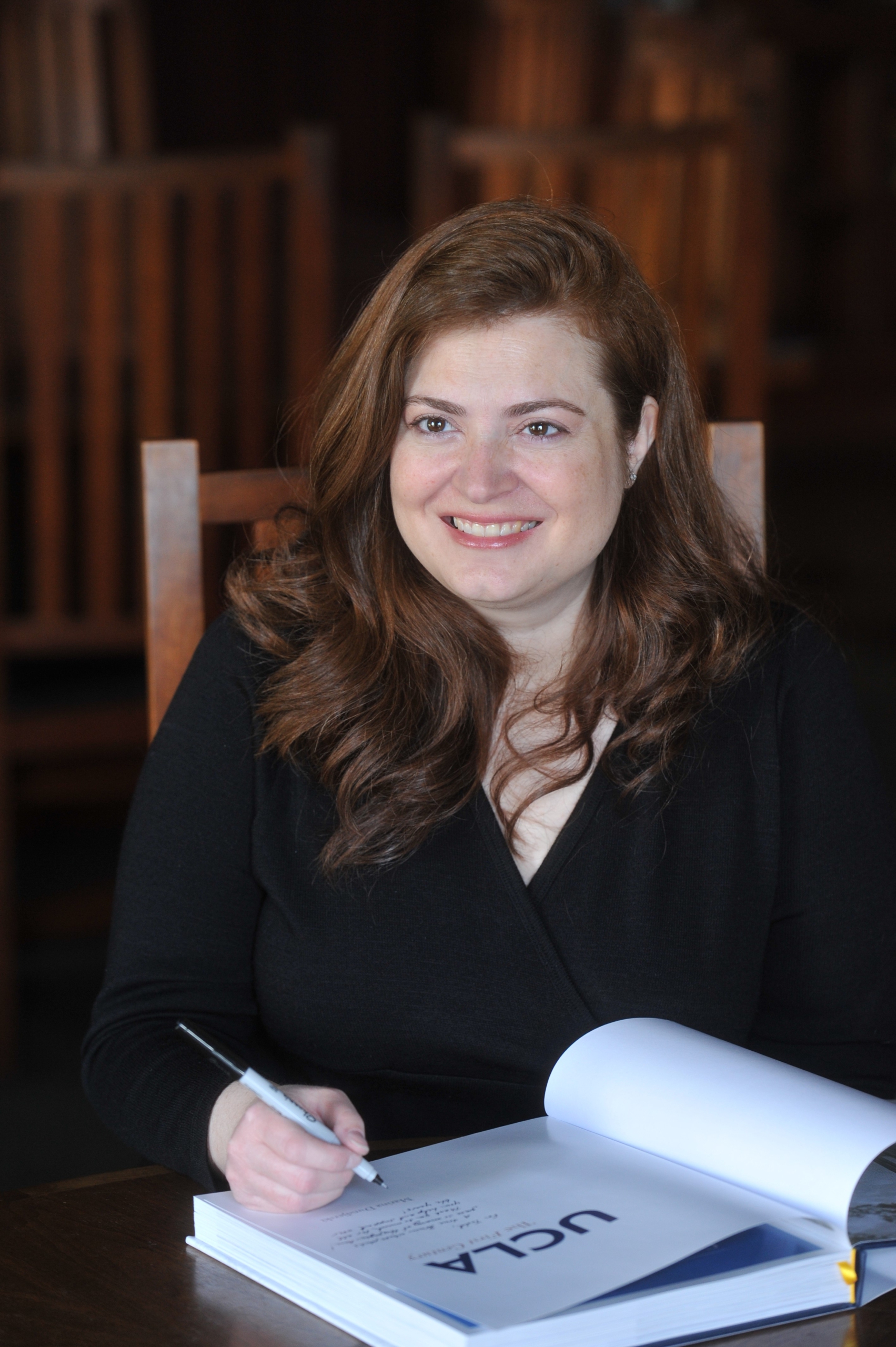 Marina Dundjerski sitting at a desk signing a copy of her book "UCLA: The First Century."