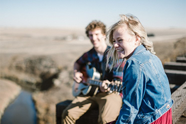 Person wearing denim jacket smiling as another person in plaid shirt strums guitar