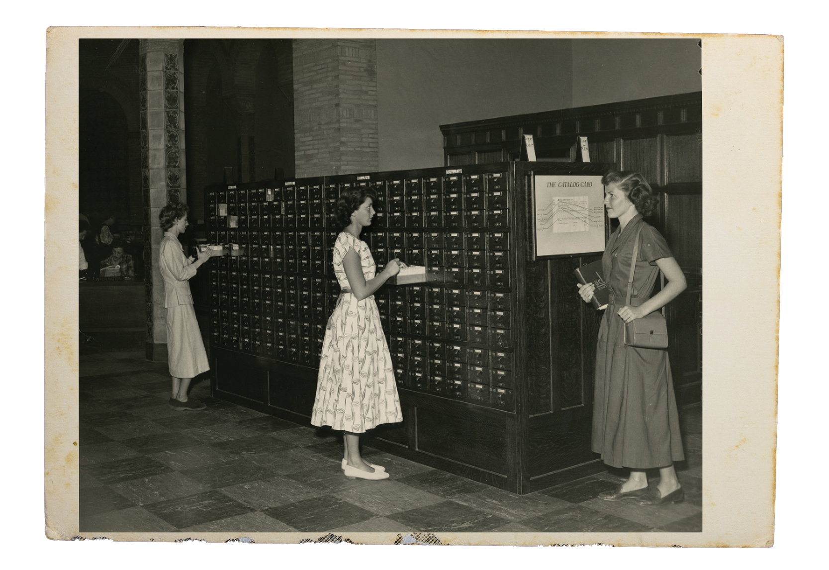 A black and white photograph of students huddled next to a library card catalog cabinet.