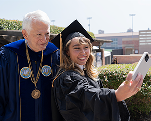 Gene Block and Katelyn Ohashi dressed in graduation regalia as they pose for a selfie.