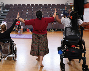 Three disabled dancers touching hands with arms raised; wheelchair user (left), person standing (center) and person standing with a walker (right)