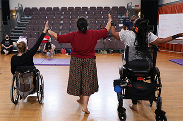 Three disabled dancers touching hands with arms raised; wheelchair user (left), person standing (center) and person standing with a walker (right)