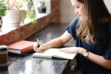 A woman wearing a blue shirt sitting a wooden table writing in a notebook.