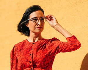 Carribean Fragoza in a red long sleeve shirt with black buttons, her left hand on her glasses as if looking at something at a distance, standing in front of a beige wall.