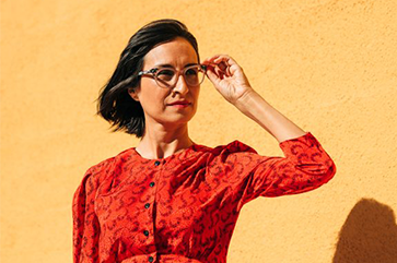Carribean Fragoza in a red long sleeve shirt with black buttons, her left hand on her glasses as if looking at something at a distance, standing in front of a beige wall.