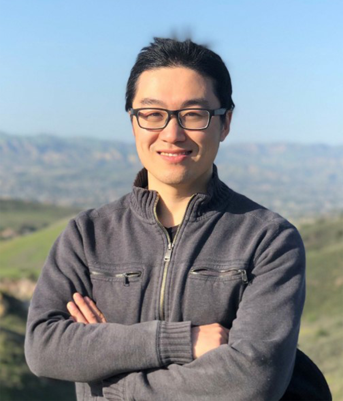 Lingsen Meng in a grey sweater with greens hills and a blue sky in the background.