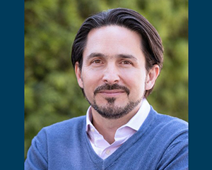 Head-and-shoulders shot of Till von Wachter in a blue v-neck sweater with a white collar shirt against a green and blurred outdoor background.