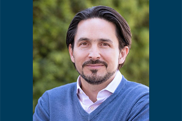 Head-and-shoulders shot of Till von Wachter in a blue v-neck sweater with a white collar shirt against a green and blurred outdoor background.